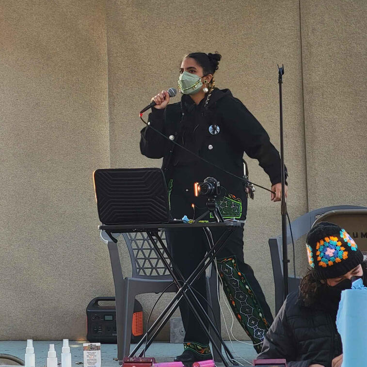 Photo from far away. Loma, a light skinned Afro-Indigenous person, is onstage in an all black outfit holding a microphone in one hand and its wire in another. They are wearing a light green N95 face mask. Sound equipment appears around them onstage.