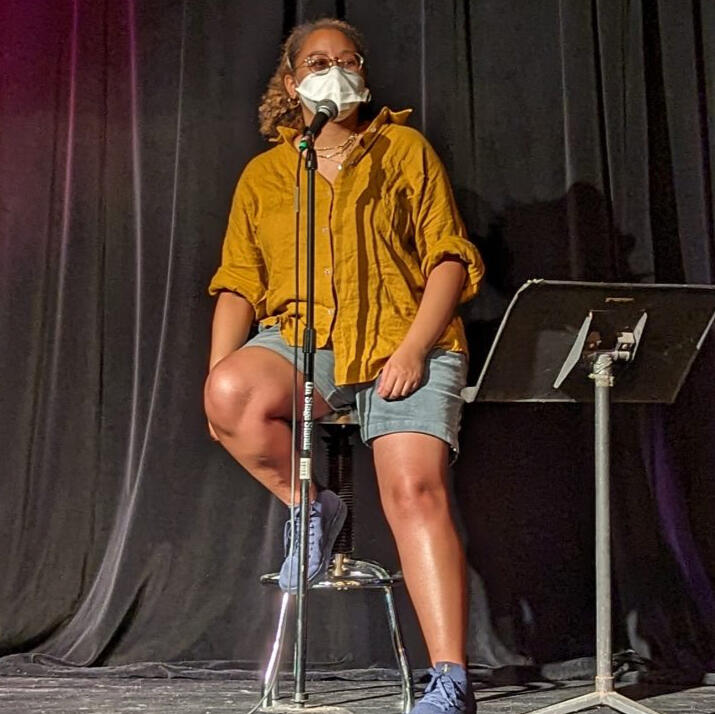 Photo. Ariana, a light skinned Black person wearing a white duckbill N95 mask, sits on a stool and speaks into a microphone on a mic stand. She is on a stage with black curtains wearing a mustard yellow shirt, blue shorts, and navy sneakers.