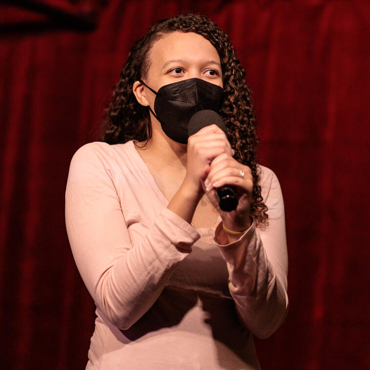 Photo. Maya, a light skinned Black person with long curly hair, is speaking into a handheld microphone they grip with both hands. They are wearing a black N95 face mask and a long sleeve light pink shirt.