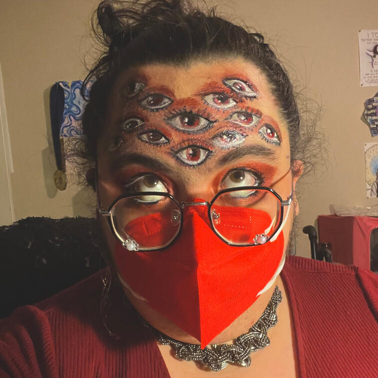 Photo. Jazz, a very light skinned person, is wearing a bright red N95 face mask, black eyeglasses, and red eyeshadow. They are looking up. Their forehead is covered in images of eyes drawn in makeup.