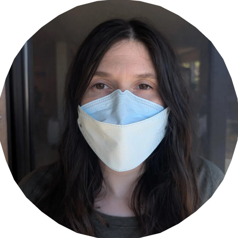 Photo. A white person with long straight dark hair with a middle part looks directly at the camera. They are wearing a white N95 face mask.